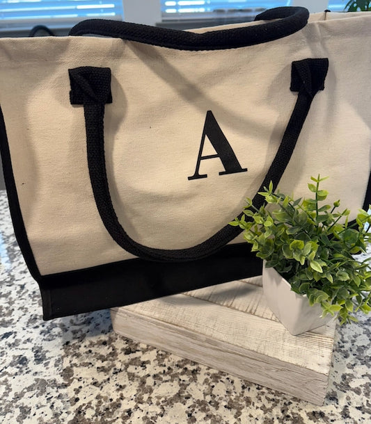 Beige tote bag with black handles and letter 'A' on a marble surface with a plant.